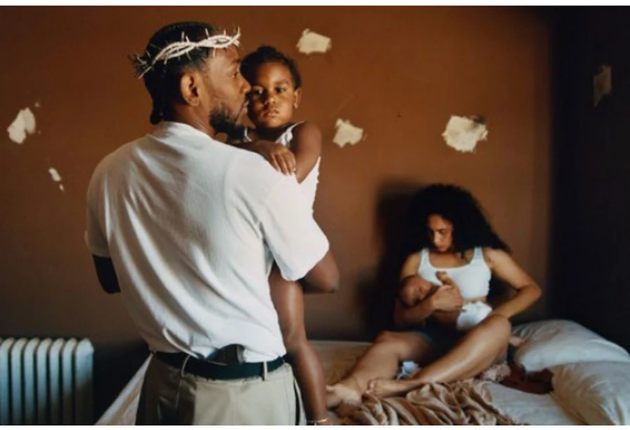 Kendrick Lamar holding a baby, with a woman seated on a bed behind him